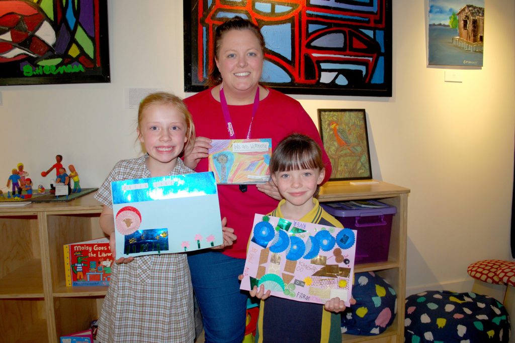 A teacher stands with two primary school girls who are showing their artwork for a book